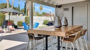 Un patio cubierto con mesa de madera y sillas de metal, sobre piso de concreto y vistas a palmeras y un jardín trasero.