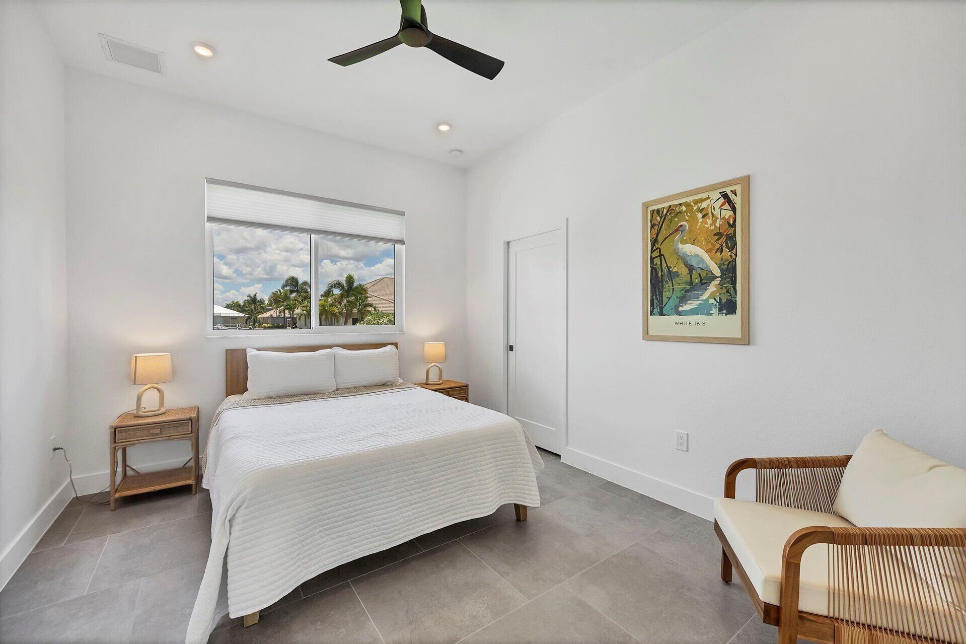 A bedroom with a bed, a window, a ceiling fan, a chair, and a painting.