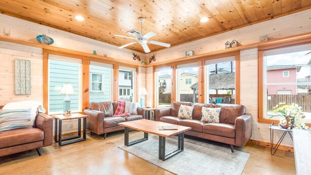 A spacious living room with a wooden ceiling, large windows, a ceiling fan, and leather furniture. - Ruby Tuesday (Port Aransas)