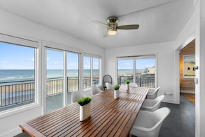 Une pièce moderne avec une table en bois, des chaises blanches et un ventilateur de plafond. De grandes fenêtres offrent une vue sur la plage.