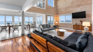 A spacious living area with a large sofa, a dining table with chairs, and a TV. The room has wooden walls and large windows offering a view of the sea. - Pool, 7 Bedrooms, Direct Oceanfront, 4 Kitchens (Port Orange)