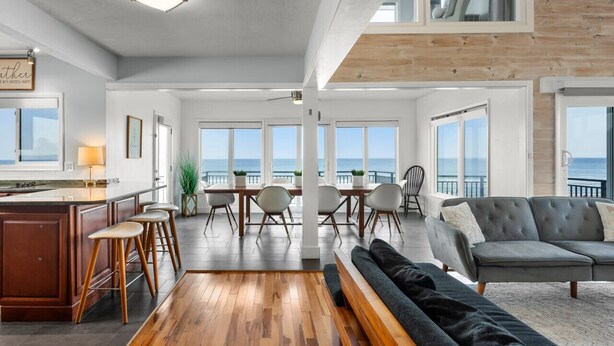 A spacious living area with a wooden floor, a grey sofa, and a dining table with chairs. - Pool, 7 Bedrooms, Direct Oceanfront, 4 Kitchens (Port Orange)