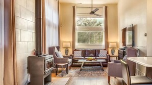 A cozy living room with a fireplace, comfortable seating, a dining area, and a large window with a view of trees. - The Addition at Villa Santorini (Gary)
