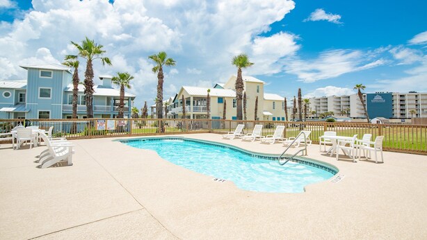 A swimming pool with lounge chairs and palm trees, surrounded by buildings with a clear blue sky. - Sunrise Villas 207 - Boardwalk TO THE Beach! (Port Aransas)