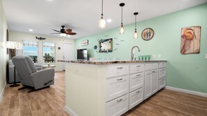A spacious living area with a large island, a gray sofa, and a ceiling fan. - Sunrise Villas 207 - Boardwalk TO THE Beach! (Port Aransas)