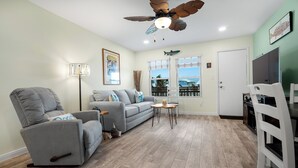 A living room with a grey sofa, a grey armchair, a small table, a floor lamp, and a ceiling fan. - Sunrise Villas 207 - Boardwalk TO THE Beach! (Port Aransas)