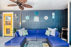 Un canapé d’angle bleu avec des coussins à motifs, une table basse en osier, un ventilateur de plafond et une porte ornée d’une pancarte « Bienvenue ».
