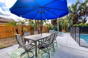 Un patio exterior con una mesa, sillas, una sombrilla azul y una piscina.