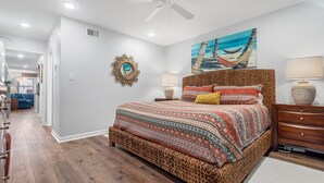 A bedroom with a large bed, a ceiling fan, a hammock painting, and a wooden nightstand. - Sunrise Villas 101 - Boardwalk TO THE Beach! (Port Aransas)