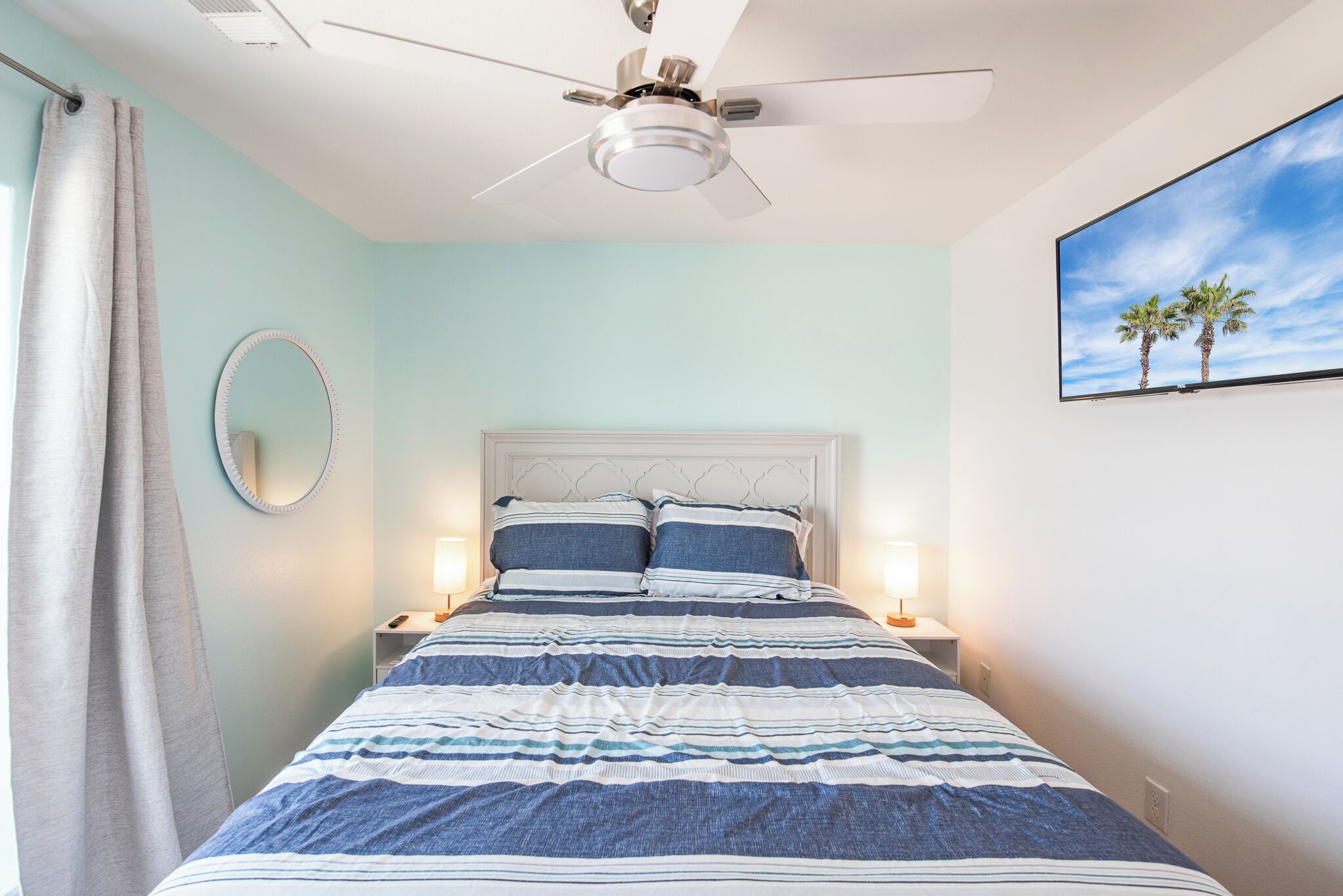 A bedroom with a bed, two bedside tables, a ceiling fan, a round mirror, and a painting of palm trees.