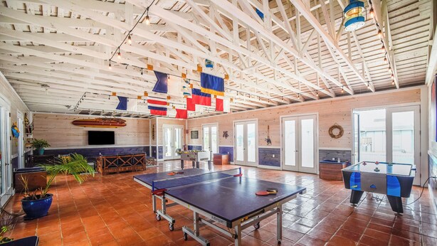 A spacious indoor area with a ping pong table, a foosball table, and a billiards table. There are potted plants and a large window providing natural light.
