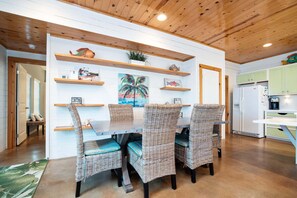 A dining area with wicker chairs, a wooden table, and a wall-mounted shelf with decorative items. - The Ritz Tarpon - Beach Gear Credit Included! (Port Aransas)