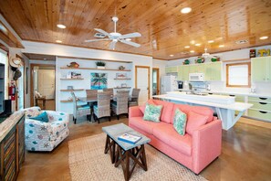 A cozy living room with a wooden ceiling, a pink sofa, wicker chairs, and a dining area with a white table. - The Ritz Tarpon - Beach Gear Credit Included! (Port Aransas)