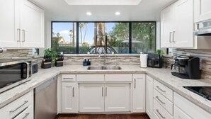 Een moderne keuken met witte kasten, een roestvrijstalen spoelbak en een groot raam met uitzicht op een zwembad en palmbomen.