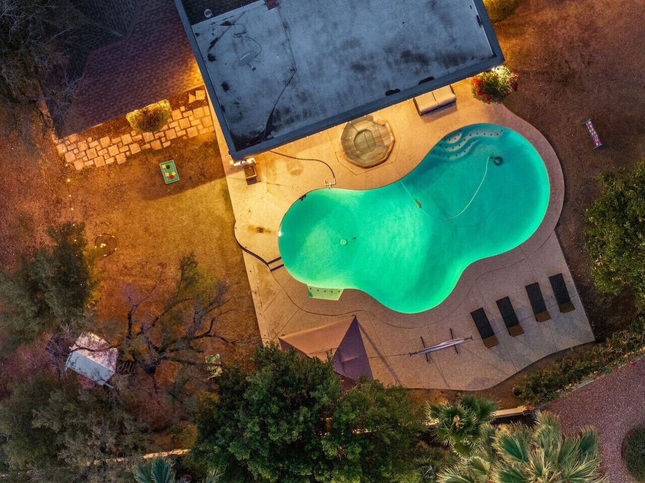 An aerial view of a swimming pool with a wavy edge, surrounded by a paved deck with lounge chairs and a small circular hot tub.