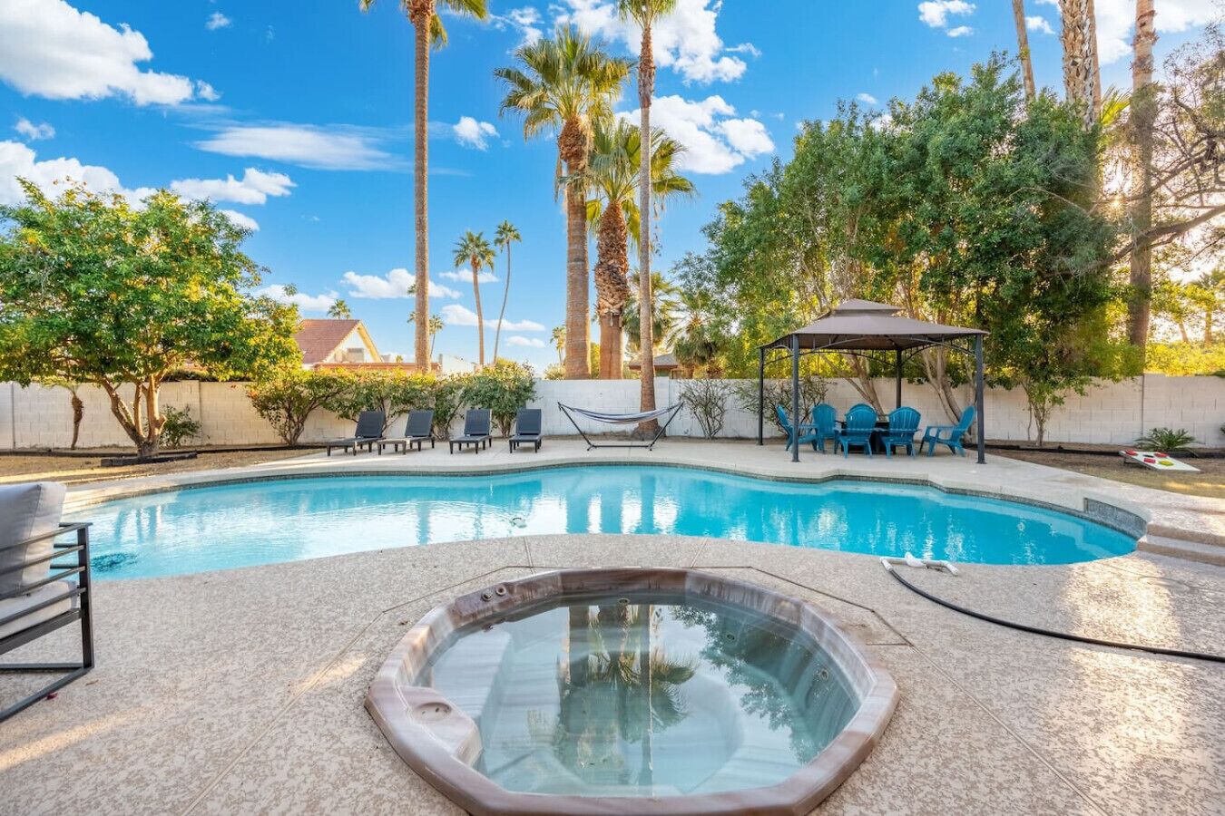 A swimming pool with lounge chairs and a hot tub, surrounded by palm trees and a covered seating area.