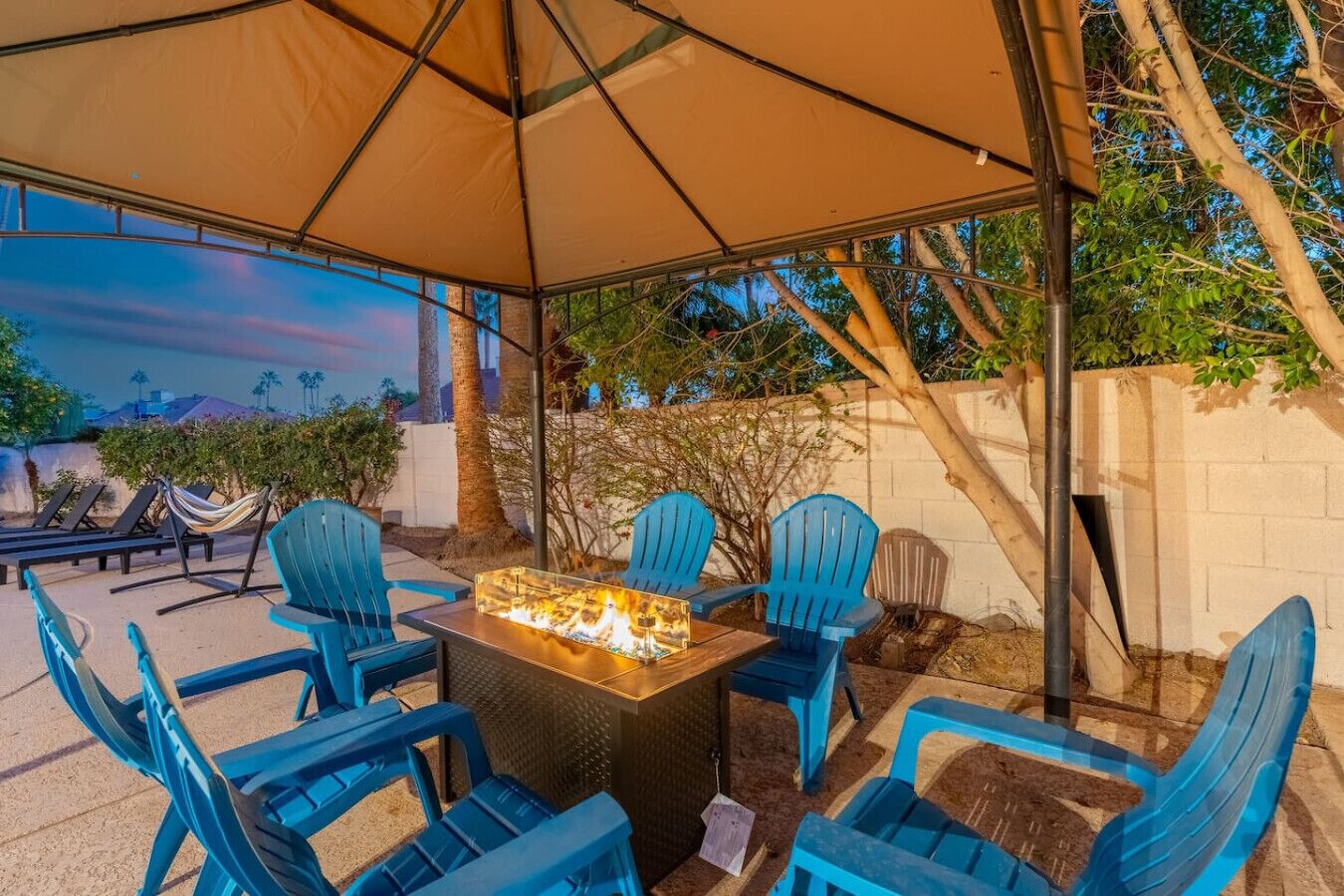 Une terrasse extérieure avec une table autour d’un foyer, entourée de chaises bleues, d’un grand parasol, et offrant une vue sur des arbres et un bâtiment en arrière-plan.