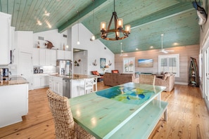 A spacious kitchen with white cabinets, a dining area with a large table, and a living room with a sofa and a TV. - Sandy Craic - Elevator! Beach Gear Credit Included (Port Aransas)