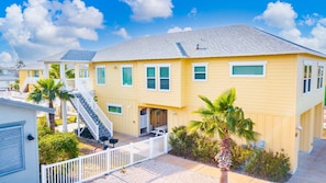 A yellow two-story house with a covered front porch, a staircase, and a fenced yard. - Sandy Craic - Elevator! Beach Gear Credit Included (Port Aransas)
