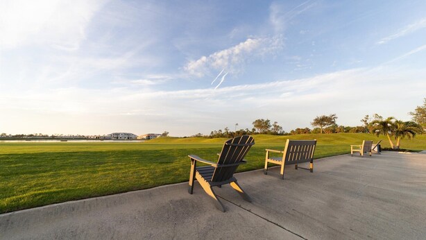 A paved walkway with two Adirondack chairs facing a grassy field and a building in the distance. - Hlb14131-828 (Punta Gorda)