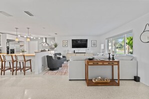 A modern living room with a white sofa, a coffee table, and a TV. There is a kitchen area with a dining table and chairs. - Sandcastle Harbor Pool & Firepit Steps to the Beach! (Ponce Inlet)