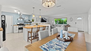 A modern kitchen with a central island, a dining area with a wooden table and chairs, and a living space with a sofa and a TV. - Sandcastle Harbor Pool & Firepit Steps to the Beach! (Ponce Inlet)