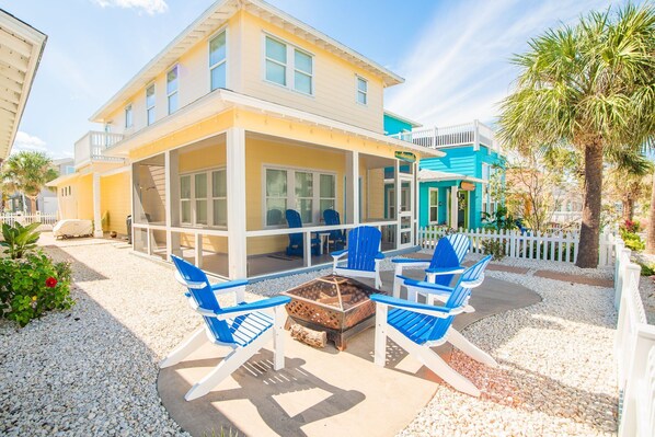 A two-story house with a covered patio area featuring blue chairs and a fire pit. - Bach Porch - Beach Gear Credit Included! 2 Shared Heated Pools! (Port Aransas)