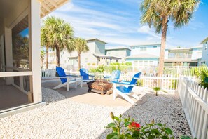 A covered outdoor seating area with blue chairs, a fire pit, and palm trees. - Bach Porch - Beach Gear Credit Included! 2 Shared Heated Pools! (Port Aransas)