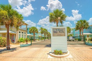 A sign for 'Village Walk' with a paved walkway, palm trees, and buildings in the background. - Bach Porch - Beach Gear Credit Included! 2 Shared Heated Pools! (Port Aransas)