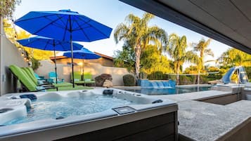 Un bain à remous entouré de pierres, deux parasols bleus et des chaises longues, le tout installé dans un jardin.