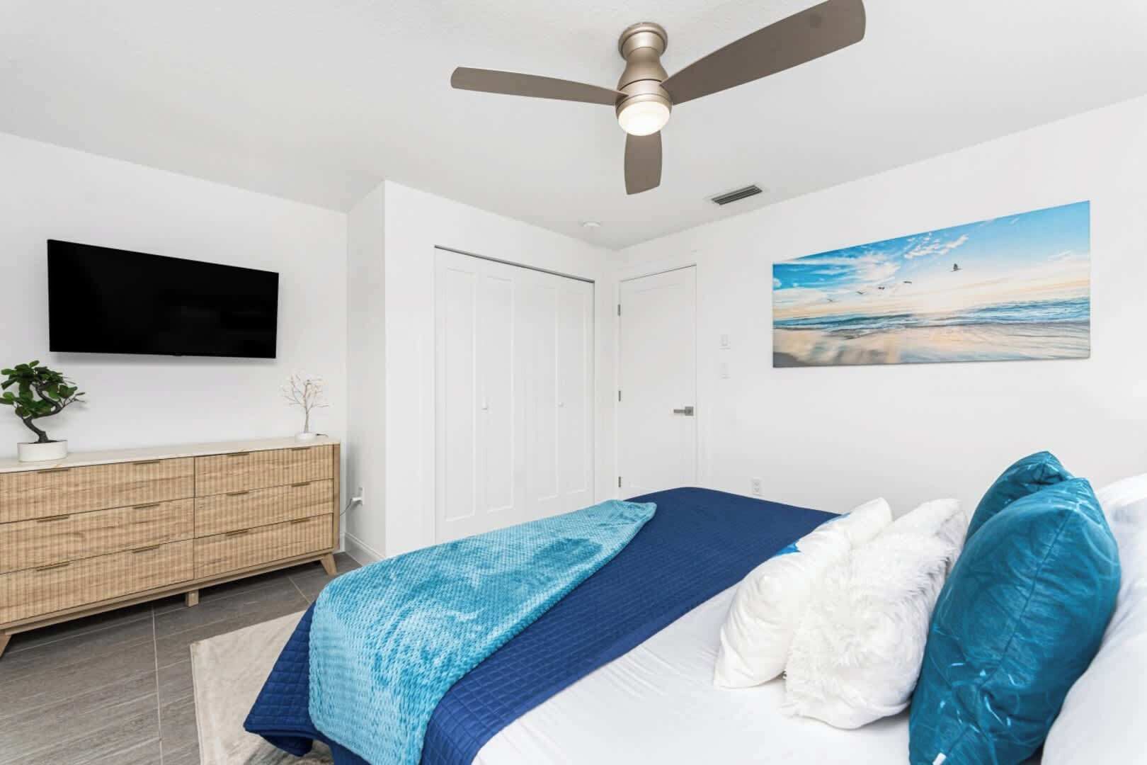 Une chambre à coucher avec un lit, un ventilateur de plafond, une télévision, une commode et un tableau accroché au mur.