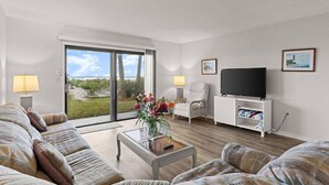 A modern living room with a sofa set, a coffee table with flowers, a TV on a stand, and a view of a garden through a sliding door. - Yellow Sunshine Oceanfront Townhouse, Private Balcony Unit #2 (Ponce Inlet)