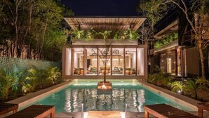 A modern pool area with a fire feature, surrounded by lush greenery and a well-lit building with large windows. - Cube House (Tulum)