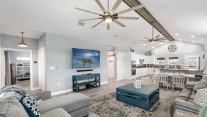A spacious living room with a large gray sectional sofa, a blue coffee table, and a ceiling fan. A kitchen with white cabinets and a dining area with white chairs is visible in the background. - Casita Del Mar - Golf Cart Included! Boat Parking! (Port Aransas)