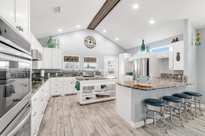 Eine moderne Küche mit weißen Schränken, einer zentralen Kochinsel mit Hockern, einer Uhr an der Wand und einem Schild, das Gäste willkommen heißt.