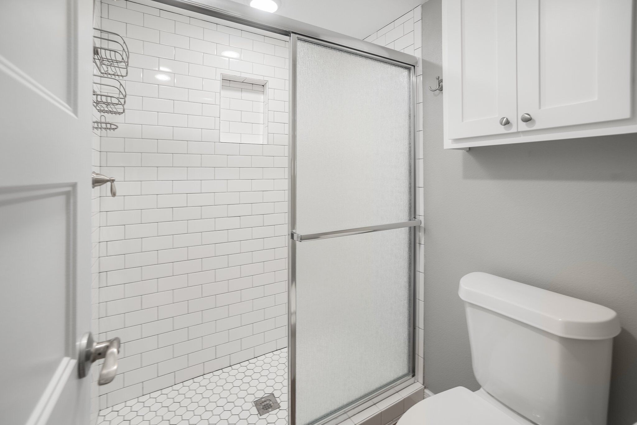 Une salle de bain avec des murs recouverts de carreaux blancs, une douche en verre, des toilettes et un meuble au-dessus des toilettes.