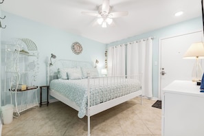 A bedroom with a white metal bed frame, a white nightstand, a white dresser, a ceiling fan, and a light fixture. - Sunrise Villa 105 - Boardwalk TO THE Beach (Port Aransas)