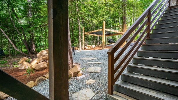 Eine Holztreppe führt zu einer Holzterrasse mit einer Bank, umgeben von bewaldetem Gelände mit Steinen und Kieswegen.