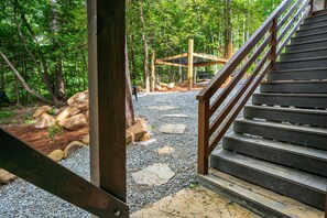 A wooden staircase leading up to a wooden deck with a bench, surrounded by a forested area with rocks and gravel paths. - Galaxy Tree House (Pittsboro)
