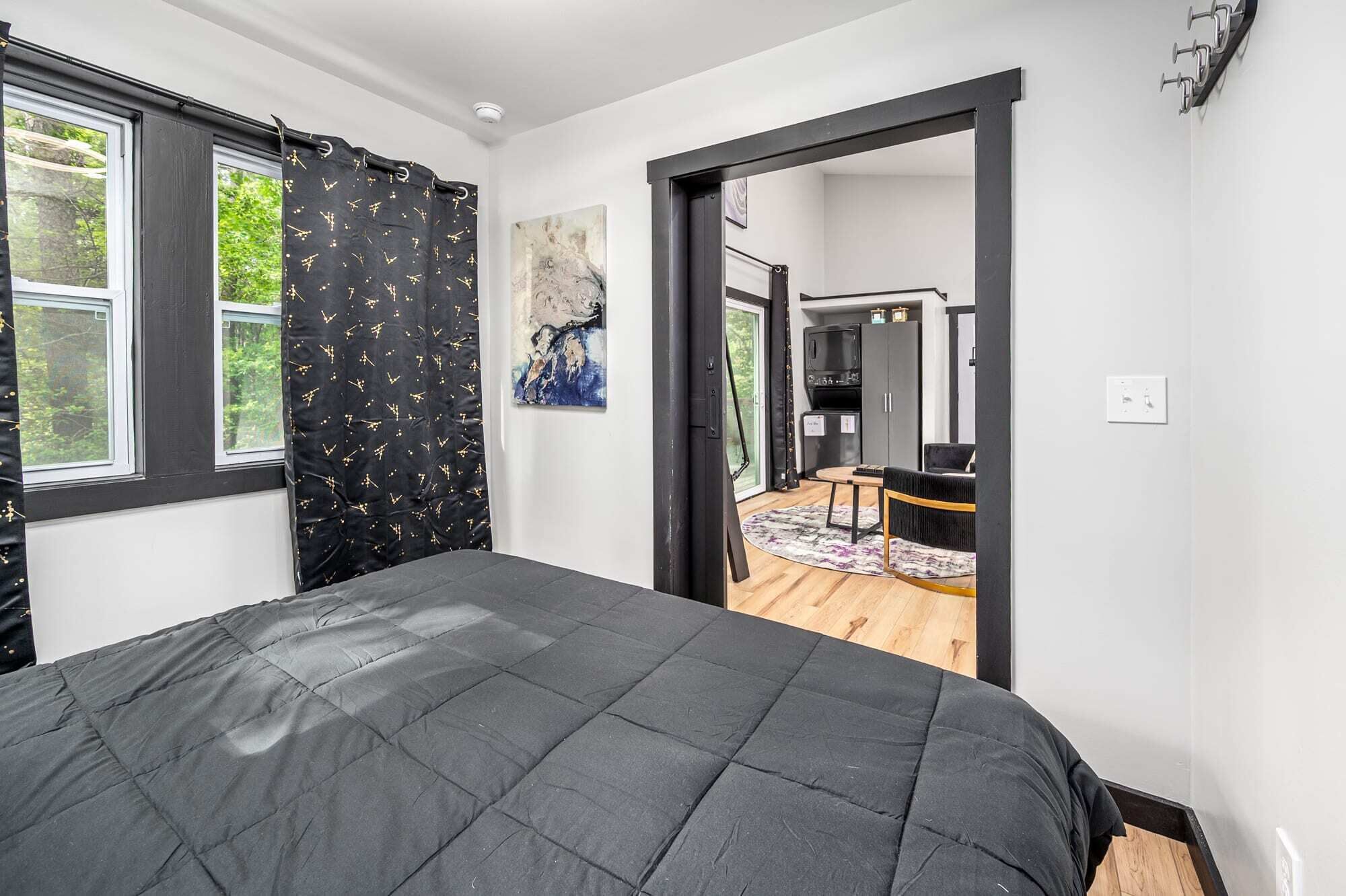 A bedroom with a large bed, a window with a black curtain, and a view of a kitchen and dining area through an open door.