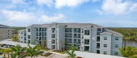 A multi-story building with balconies, a parking area, and palm trees.