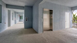 Un couloir avec un ascenseur et une porte, qui mène à un balcon d’où l’on peut admirer les voitures garées et les palmiers.