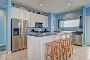 A kitchen with white cabinets, a stainless steel refrigerator, a microwave, and a dishwasher. There are three wooden stools at the island. - Casa Las Palmas - Boardwalk to the Beach! (Port Aransas)