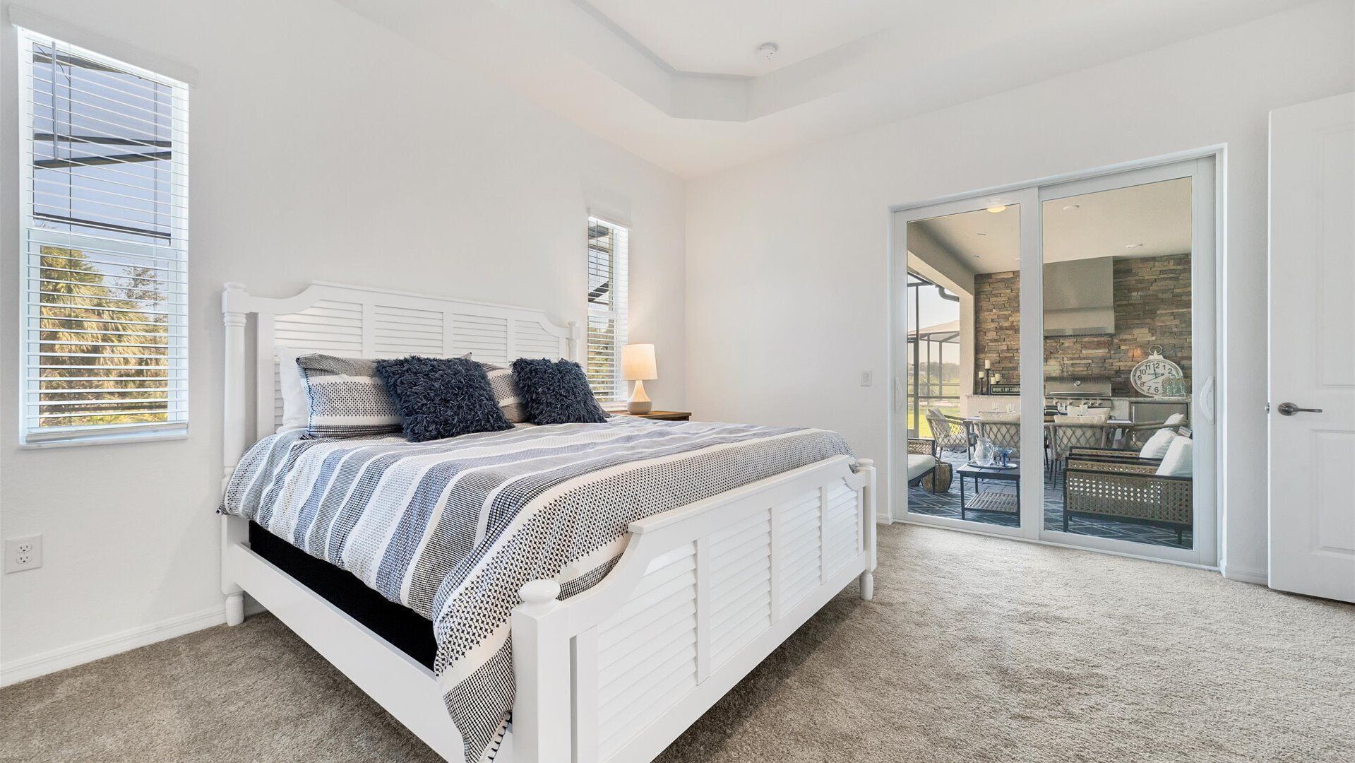 Ein Schlafzimmer mit einem weißen Bett, gestreifter Bettwäsche und zwei Kissen. Es gibt ein Fenster mit Jalousien, eine Tür zu einem Balkon und einen Blick auf eine Außenpatio-Terrasse.