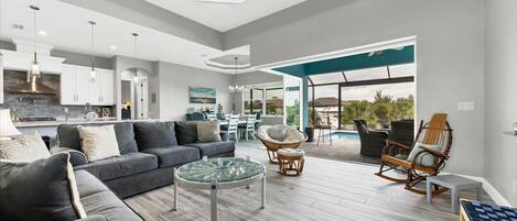 A modern living room with a grey sofa, a glass coffee table, wicker chairs, and a view of a pool and outdoor area.