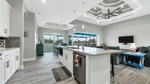 A modern kitchen with a large island, a dining area with chairs, a blue accent wall, and a TV mounted on the wall. - Mpc15528 (Port Charlotte)