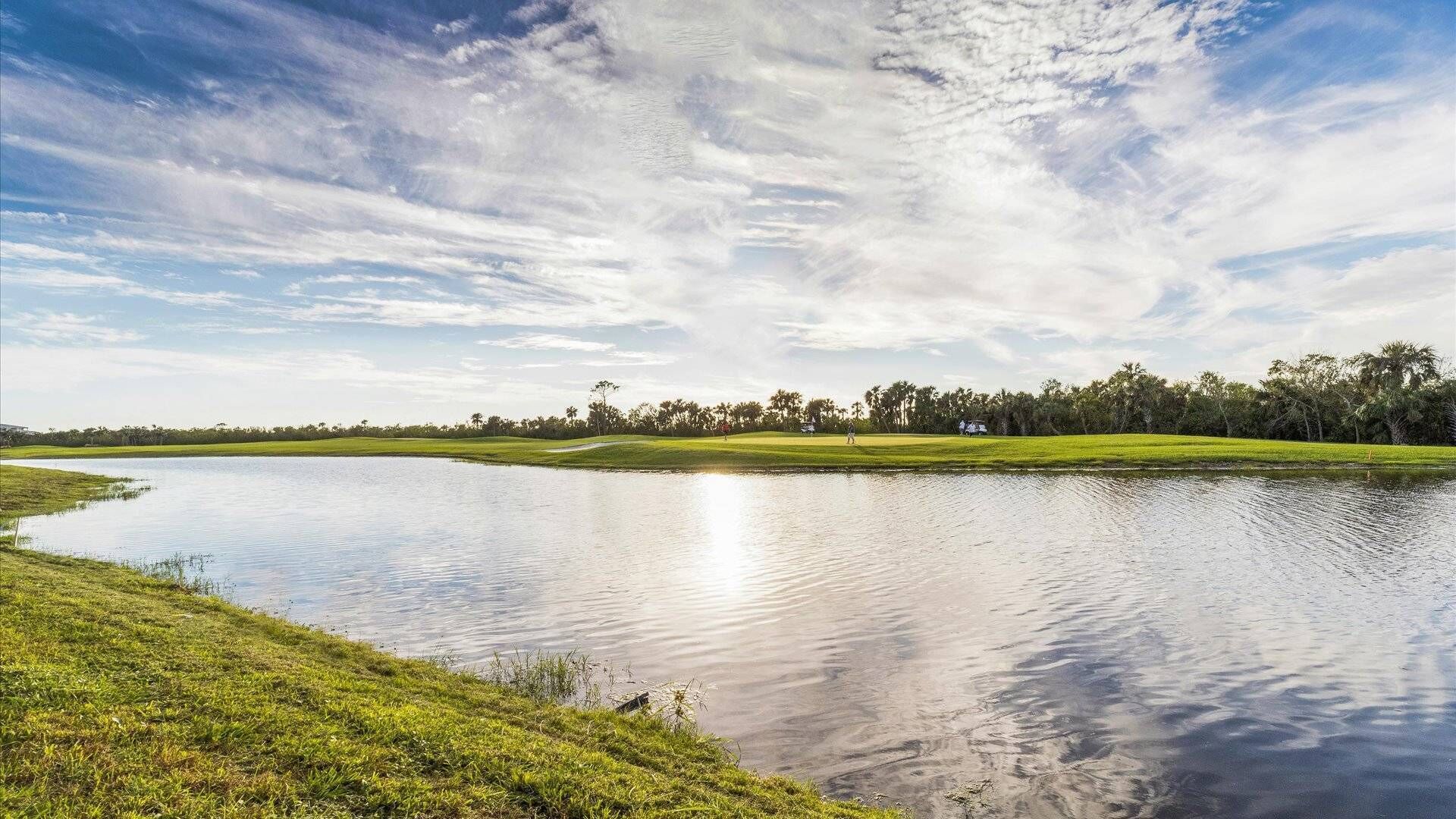 Un paisaje sereno con un lago tranquilo, hierba verde y una hilera de árboles bajo un cielo parcialmente nublado.