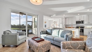 A spacious living room with a large window, a grey sofa, patterned ottoman, and a dining area with a kitchen in the background. - Hlb14041-613 (Punta Gorda)