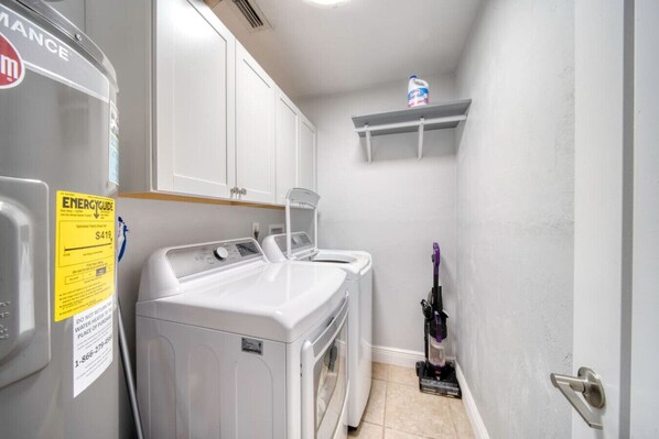 Une buanderie équipée de deux machines à laver et deux sèche-linge à chargement frontal, d’un aspirateur et d’une étagère sur laquelle se trouve une bouteille de lessive.