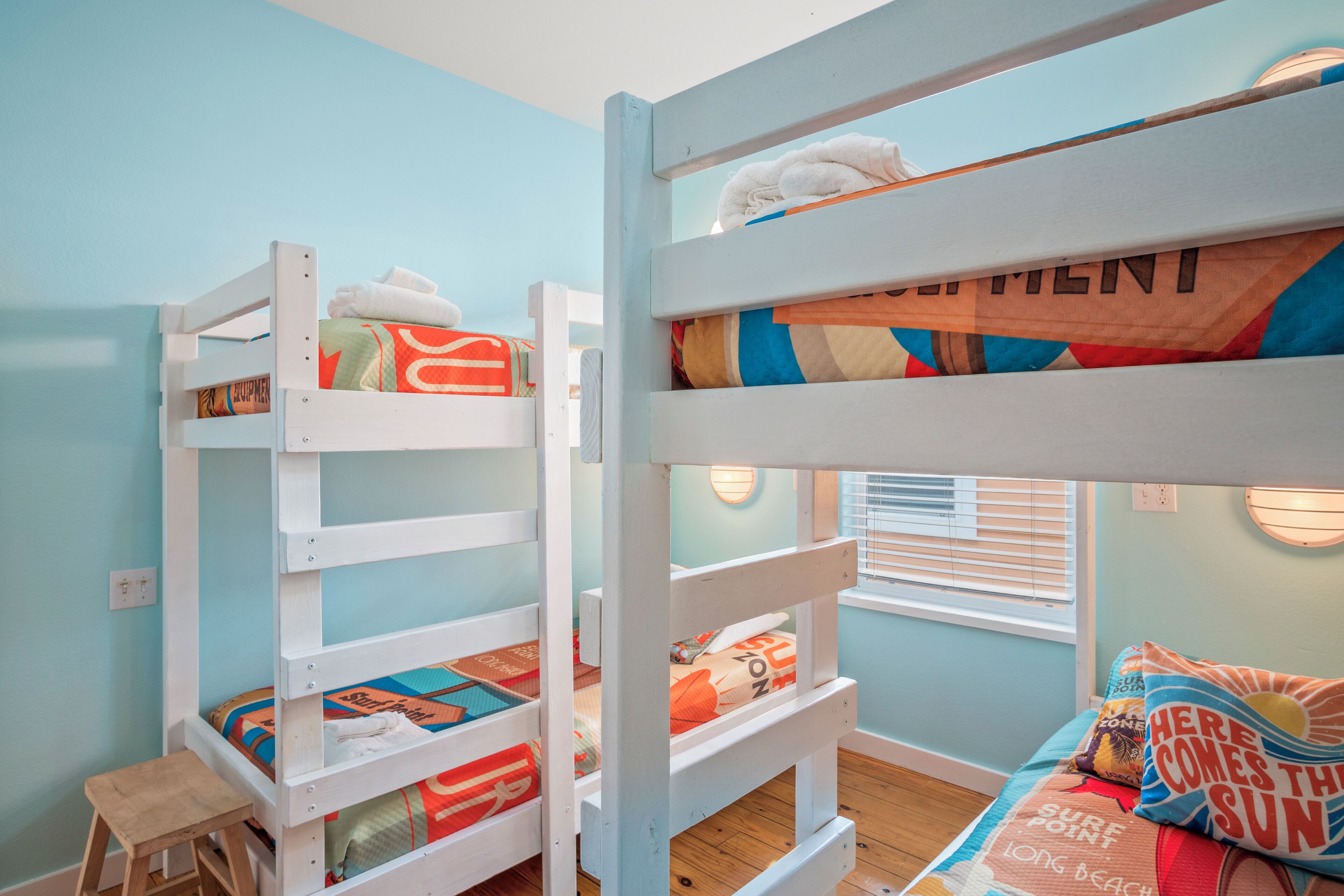 A bunk bed room with a small stool, a window with blinds, and a wall-mounted light fixture.